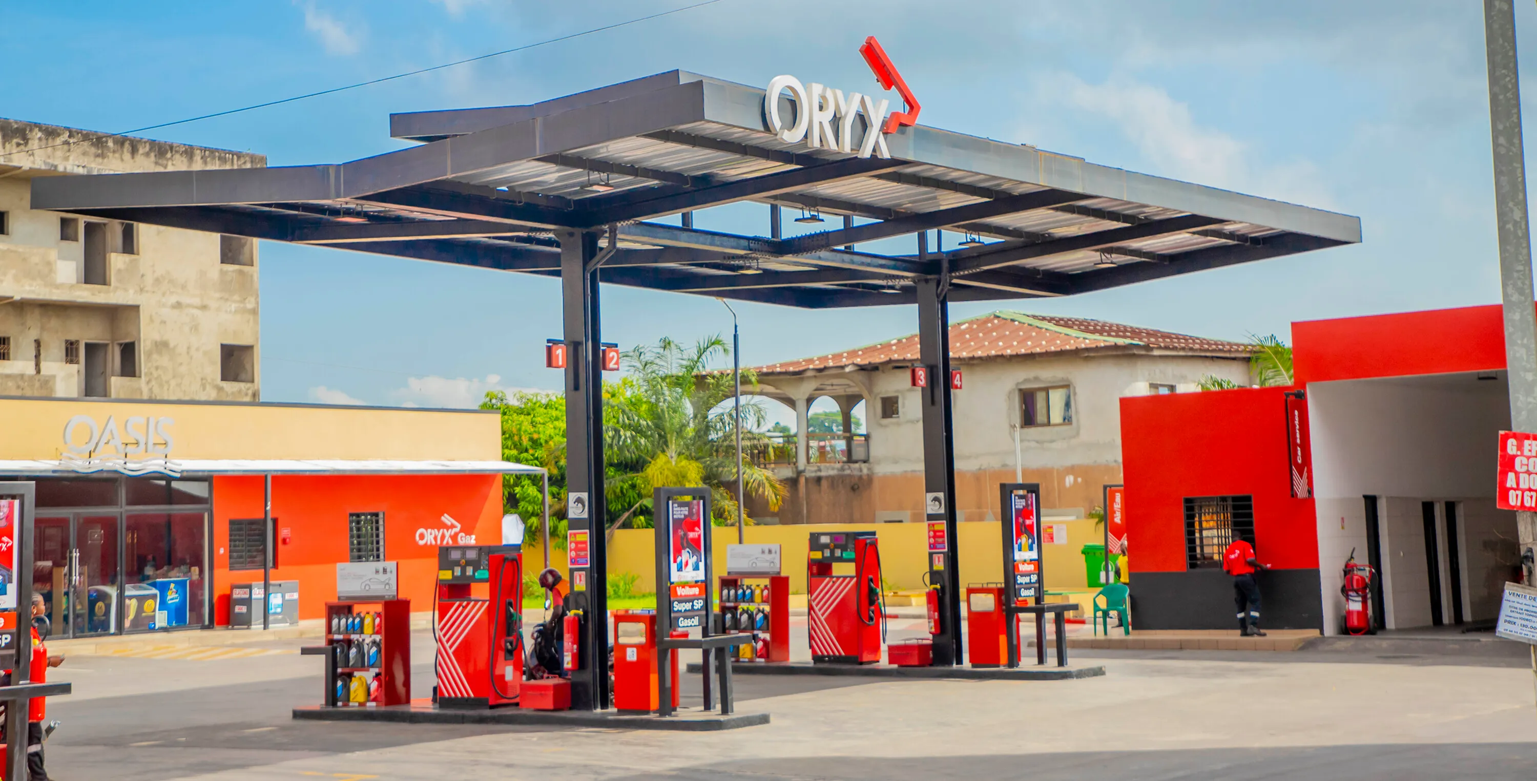 Modern fuel station with red and black pumps, convenience store in background, blue sky above.