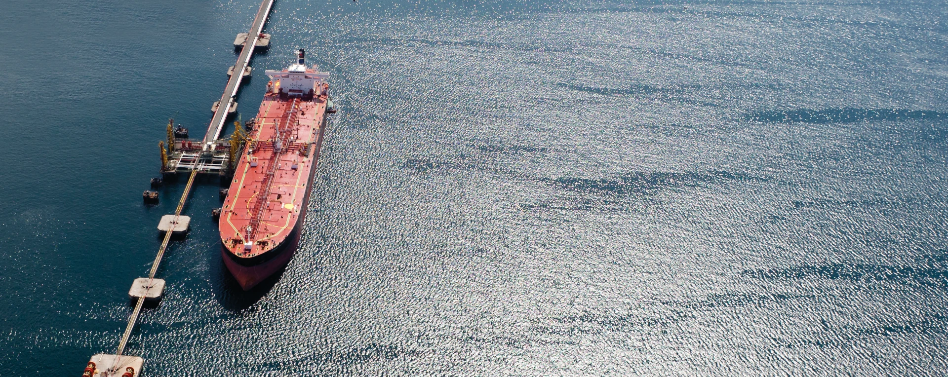 Large red cargo ship docked at a pier in calm, sunlit water.